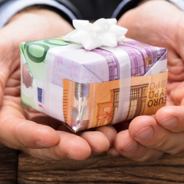 Hands cupping a small gift box wrapped in euro banknote–themed paper with a white bow on a wooden table.